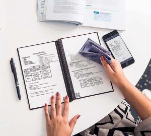 Table top with a person wiping off what they'd written on the Rocketbook Fushion. Cell phone next to the jounral shows that what was written in the journal has transferred to the phone.